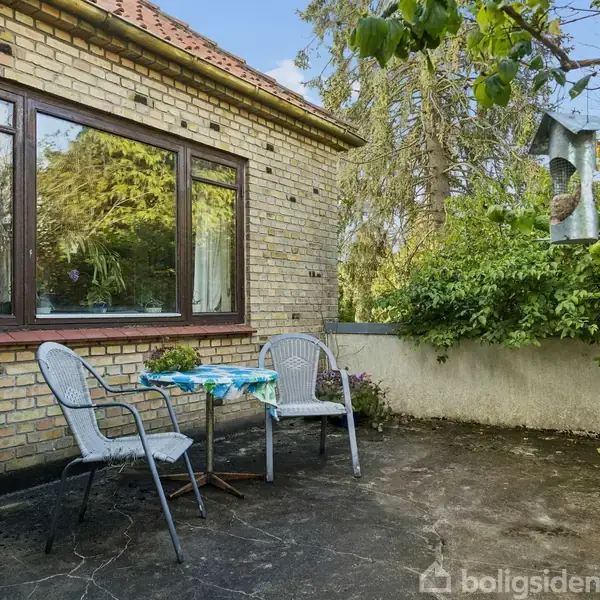 Et lille bord med to stole står på en terrasse af beton, op ad en husmur af gule mursten. Omgivet af grønne træer og buske.
