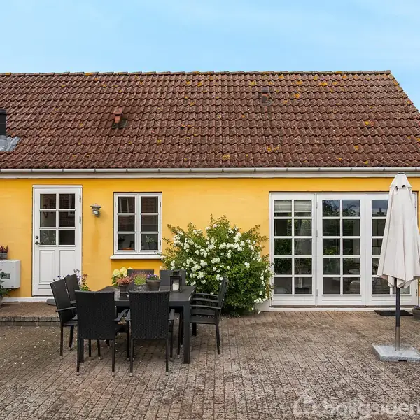Et gult hus med rødt tegltag, hvor en terrasse foran har sort udemøblement og en parasol. Døre og vinduer åbner mod terrassen, omgivet af blomster og buske.