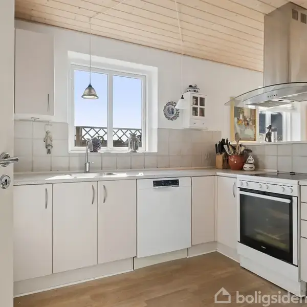 Lyse, moderne køkken med hvide skabe og skuffer, indbygget ovn og opvaskemaskine. En vask med vindue ovenfor, hængende lampe, og køkkengrej på bordpladen. Trægulv og lofter skaber en hyggelig atmosfære.