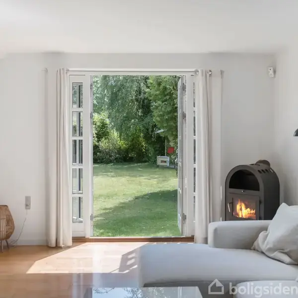 En stue med en lys sofa, pejs tændt, åben dør fører ud til en grøn have. Et hvidt gardin hænger ved døren, skabsarrangement til venstre.