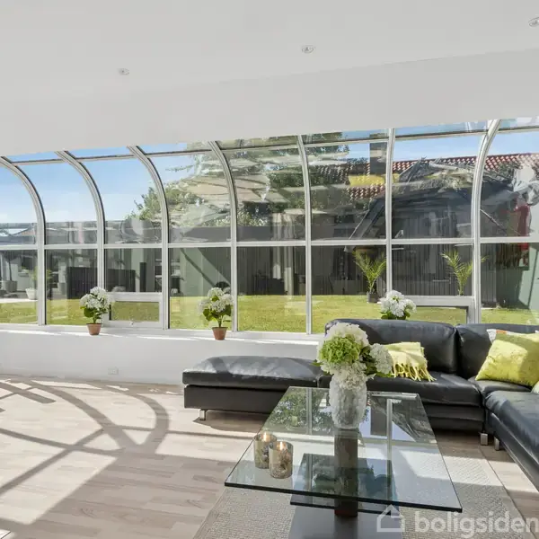 En moderne stue med sort lædersofa og grønne puder, der står foran et stort buet vindue. Udsigt til en have udenfor. Glasbord med blomster i midten af rummet.