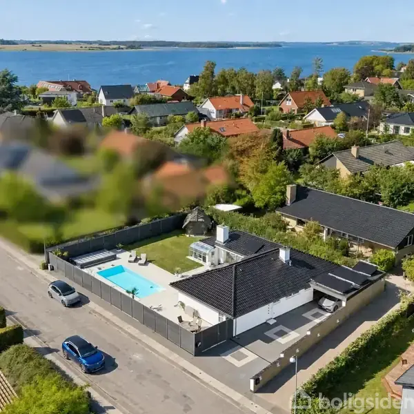 Huset er omgivet af en hæk og har en stor terrasse med en swimmingpool. Biler er parkeret på vejen foran huset. I baggrunden ses boligkvarter og vandudsigt.