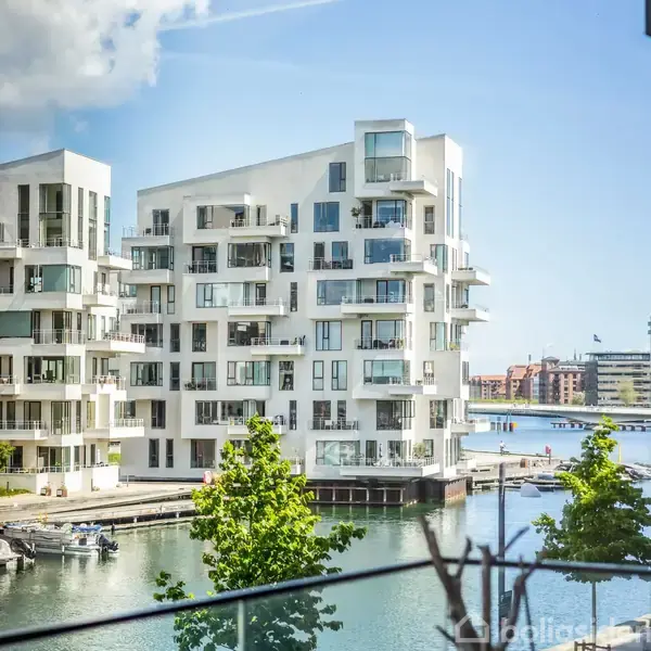 Hvide moderne lejlighedskomplekser står ved kanten af en vandkanal. Solen skinner kraftigt, og flere både er fortøjet langs kajen. Små træer ses i forgrunden.