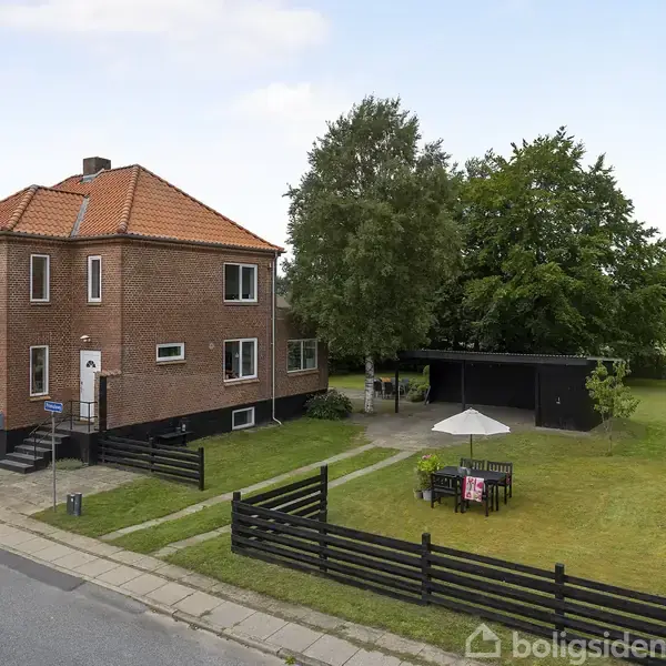 Et rødt murstenshus med orange tag står ved en vej. Foran er en have med et hvidt parasol over et bord og stole, omgivet af græs og træer.