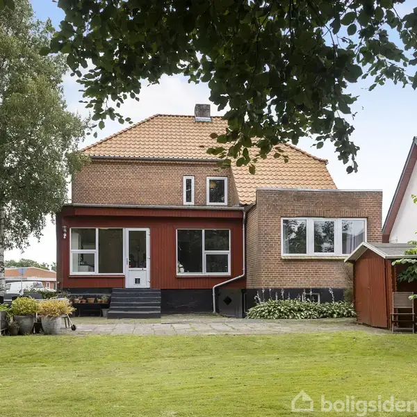 Hus i røde mursten med gavltag, store vinduer og en have foran. Flisebelagt terrasse og skur til højre. Grønne træer omkranser området. Boligsiden-vandmærke nederst.
