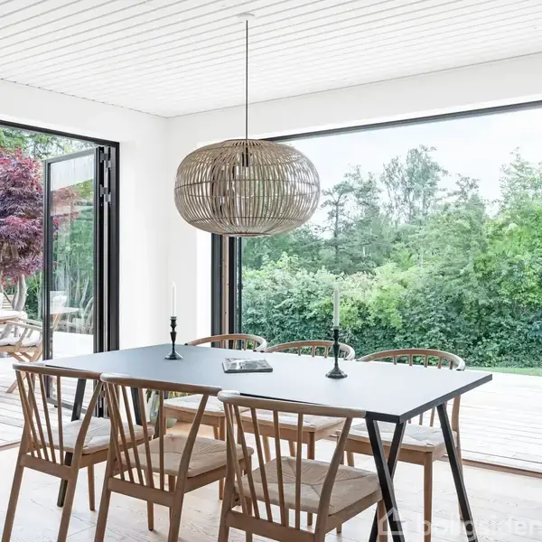 Spisebord med træstole står i centrum, indrammet af store vinduer, der åbner mod en terrasse med grøn udsigt og rødligt træ. En stor, flettet lampe hænger over bordet.