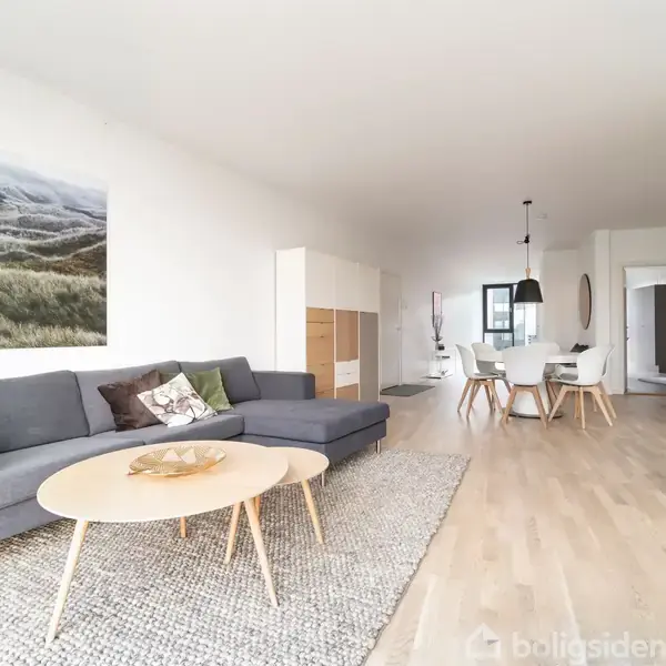 Grå sofa står stille i en lys stue med en plakat af landskab på væggen, træbord foran, spisebord i baggrunden, lyse trægulve og dekorative planter.