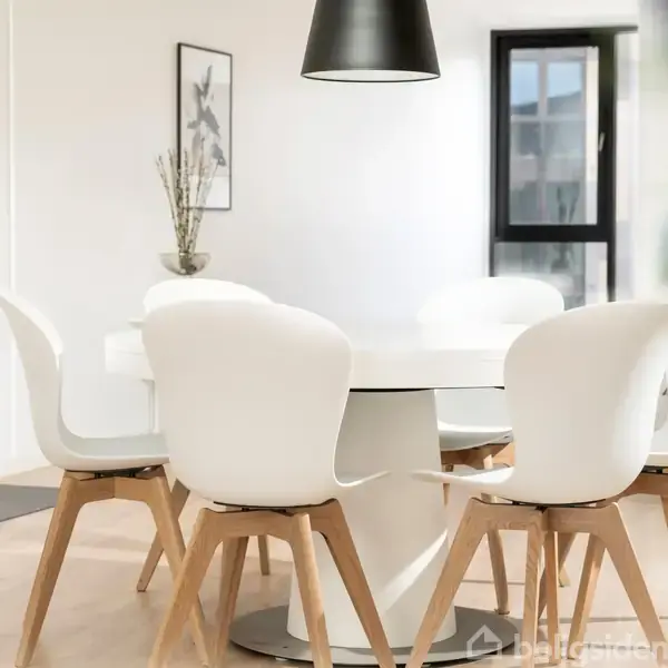 Et rundt, hvidt spisebord med seks hvide stole omkring det, står i en moderne spisestue. En sort loftslampe hænger over bordet, og der er et vindue i baggrunden.