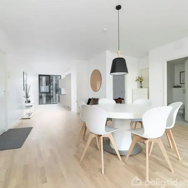 Rundt spisebord med fem hvide stole står i en moderne, lys stue med trægulv. En sort lampe hænger over bordet, og en stor vægdekoration ses i baggrunden.