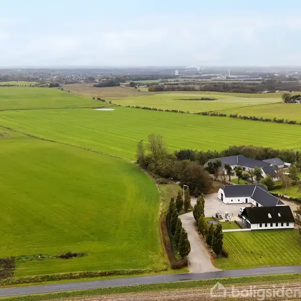 Hvidt hus står stille omgivet af store grønne marker og spredte træer i et åbent landligt område med en vej i forgrunden og flere bygninger i det fjerne.