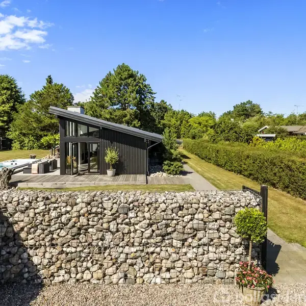 Et moderne sort-hus med store vinduer står i en grøn have med svømmebassin. Stenmur og åben port markerer indgang. Himmel er klar og blå med få skyer.