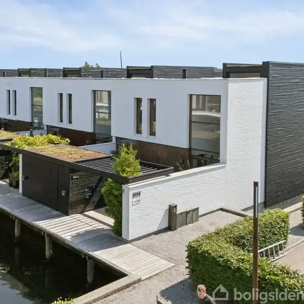 Rækkehus med moderne design og fladt tag står ved en kanal. Terrasser har grønne planter. Husklyngen omgives af hække og flisebelagte stier.