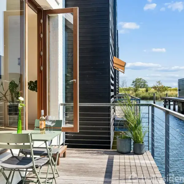 Balkon med bord og stole står ud mod vandet. En åben dør fører til indendørs, hvor planter er synlige. Omgivelserne inkluderer flere bygninger og en blå himmel med skyer.