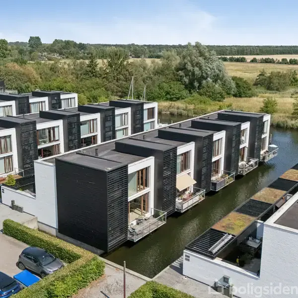 Rækkehuse ved en kanal med moderne sort-hvid arkitektur. Bygningerne står langs vandet med altaner, omgivet af parkeringsplads med biler og grønne områder i baggrunden.
