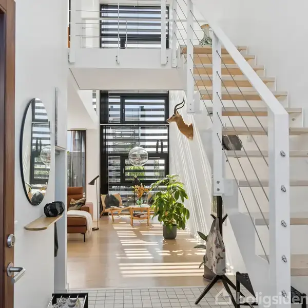 Trappeopgang fører op til øverste etage i moderne, lyst hus. I midten står en stor grøn plante, omgivet af stole, mens sollys strømmer ind gennem store vinduer.