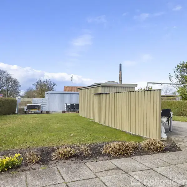 Græsplæne omgivet af lave buske. Et plankeværk og terrasse med havemøbler til højre. Baghus og hegnet område i baggrunden. Klar blå himmel.