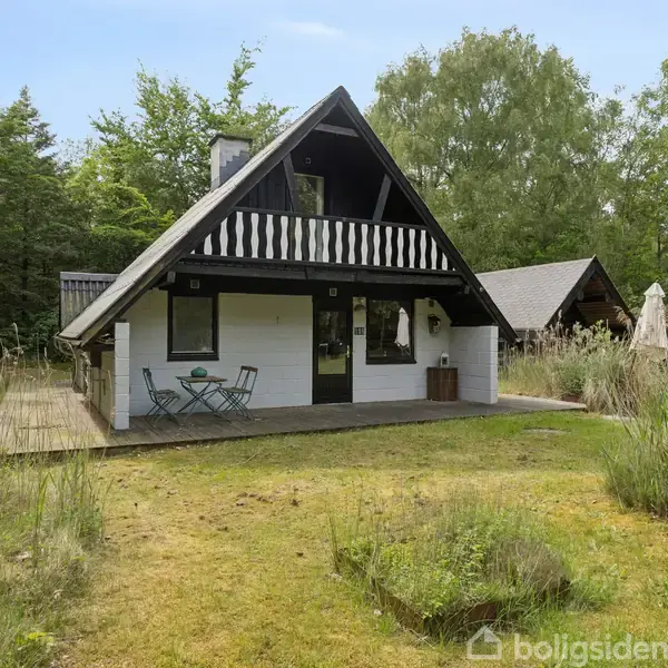 Et sommerhus står stille i en skovklædt have. Foran huset står et lille bord og to stole på en træterrasse. Omkring huset er tæt vegetation og træer.