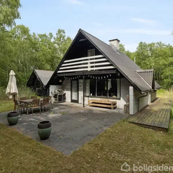 Triangulært sommerhus med sort træfacade og store vinduer, står på en flisebelagt terrasse med havemøbler og grill, omgivet af frodig skov og græsplæne.