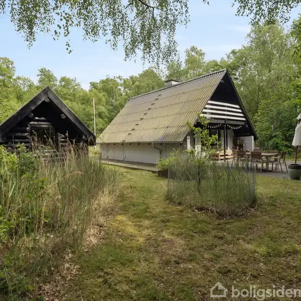 To huse står på en græsplæne omgivet af skov. Det ene hus har en udendørs terrasse med borde og stole samt en parasol ved siden af.