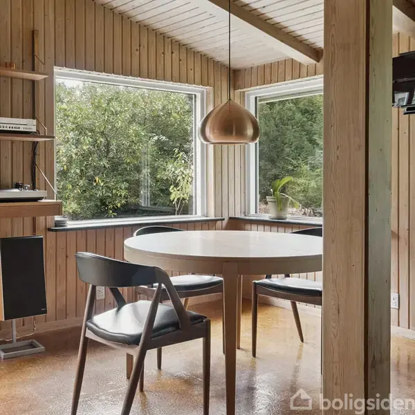 Rundt spisebord med tre sorte stole står i et hjørne af et træbeklædt rum. En kobberlampe hænger over bordet. To store vinduer viser frodig udendørs vegetation.