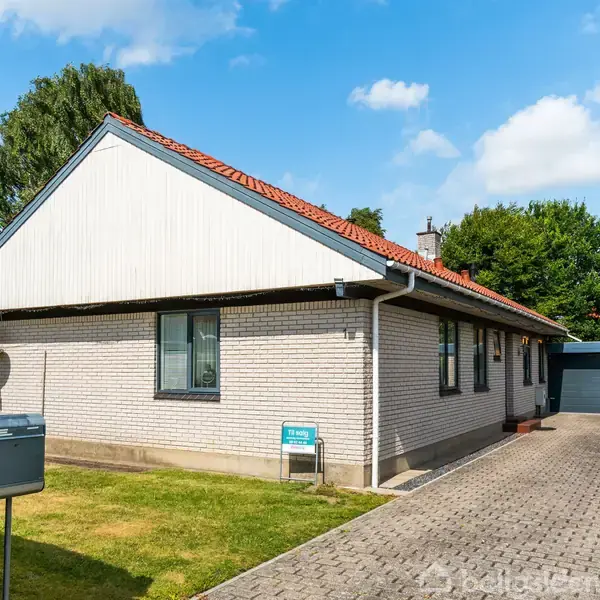 En lys murstensvilla står stille omgivet af en velholdt have med grønne buske. Der er en flisebelagt indkørsel ved siden af huset. Skyfri blå himmel i baggrunden.