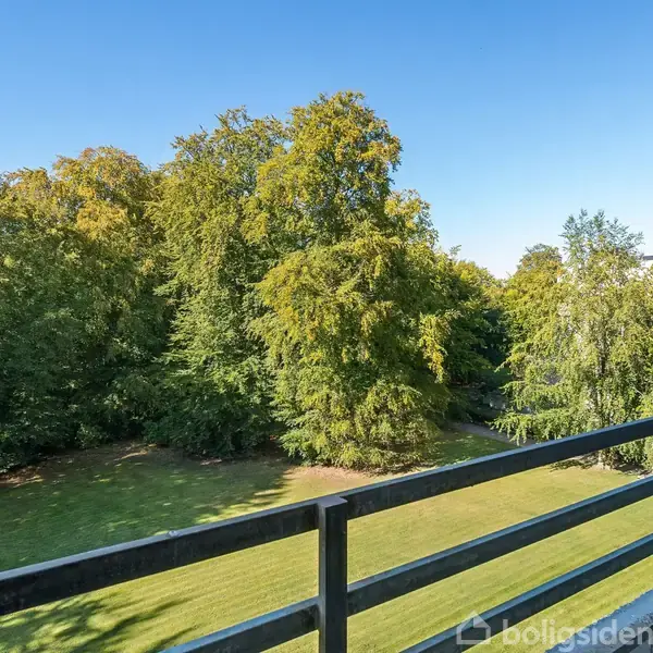 Balkon med udsigt over en grøn park fyldt med store træer. I baggrunden ses en moderne bygning under en klar blå himmel.