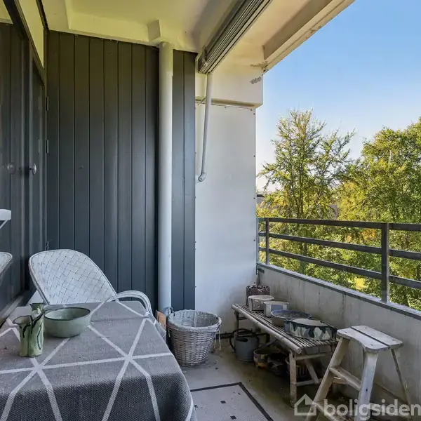 En balkon med en hvid stol og et tæppebeklædt bord, omgivet af grønne træer i baggrunden. En lille hylde og kurv står ved siden af, hvilket skaber en hyggelig atmosfære.