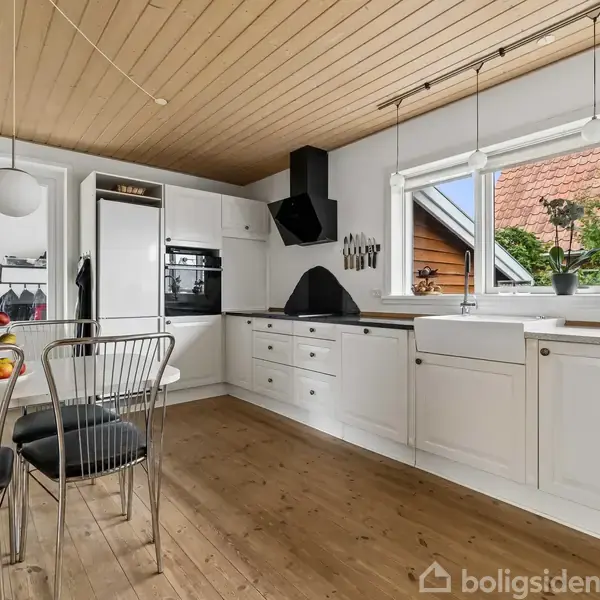 Et rødt murstenshus med en skråtag er omgivet af en have. Der er en veranda på siden og en lille balkon. Himlen er blå med spredte skyer.