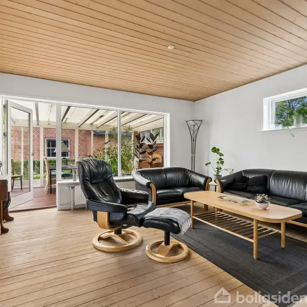 Et rødt murstenshus med spidst tag står i en frodig have. Foran huset er der en græsplæne og buske. Nabohuse kan ses i baggrunden under en blå himmel.