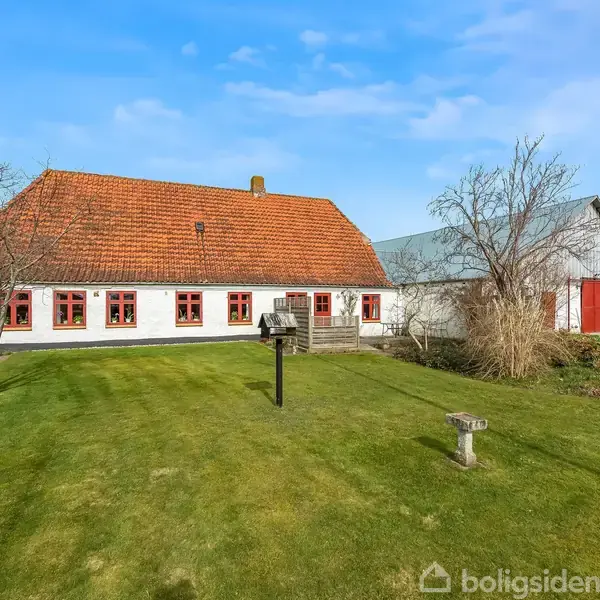 Hus med hvid facade og rødt tag står i en velplejet have. Der er redskabsskur ved siden af huset. Græsplænen omringes af buske og træer, under en klar blå himmel.