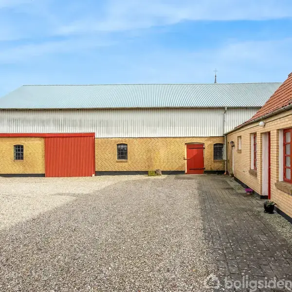 Gårdbygning med gul murstensfacade og røde døre, der står stille i en grusbelagt gårdsplads, omkranset af en blå himmel.