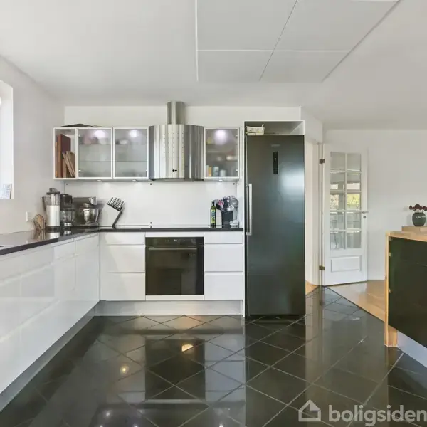 Køkken med hvide skabe og sorte bordplader, udstyret med sort komfur og emhætte. Højskab ved siden af glasdør. Vindue giver udsigt til træer. Mørke gulvfliser.