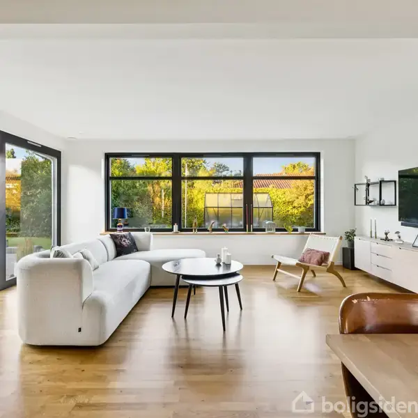 En hvid sofa står i en lys stue med trægulv, omkranset af store vinduer, som viser en grøn have udenfor. Foran sofaen er der et rundt sofabord.