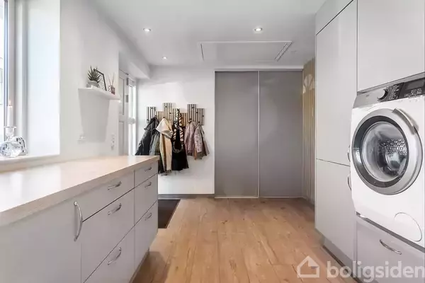 Vaskemaskine installeret i en moderne bryggers med lyst trægulv; skabe langs væggen til opbevaring. Flere jakker hænger på knager ved væggen. Lys strømmer ind fra vinduerne.