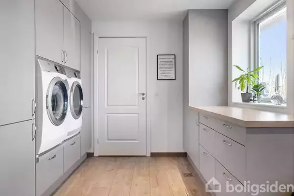 Vaskemaskine og tørretumbler er indbygget i en grå skabsvæg, placeret i et lyst bryggers med trægulv. Et vindue med en plante står til højre for indgangen.