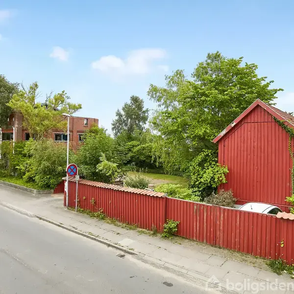 Et rødt hus står stille omgivet af frodige træer og buske, ved siden af en vej. Et skilt med teksten "C 11 3" er placeret tæt på hegnet.