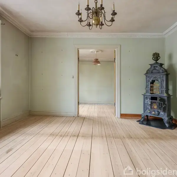 En antik kamin står stille i en lys stue med trægulv og grønne vægge. En lysekrone hænger fra loftet, og et vindue og en døråbning ses i baggrunden.