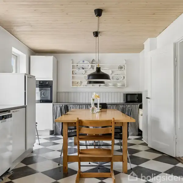 Et træbord med stole står centralt i et køkken med sort-hvid ternet gulv, hængende lampe, og åbne hylder med porcelæn. Rummet er lyst med træloft og en analog urskive.