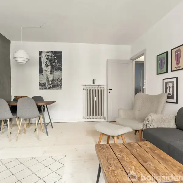 Sofabord i træ står foran en grå sofa i en stue med hvidmalede vægge. Spisebord med fire stole ved siden af en radiatorskulptur. Billeder hænger på væggene.