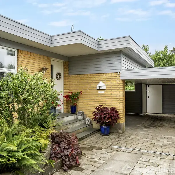 Hus indgang med mursten og træbeklædning, omgivet af planter og blomster i krukker på trappe. Carport til højre, flisebelagt indgangsparti. Husnummer over døren: 95.