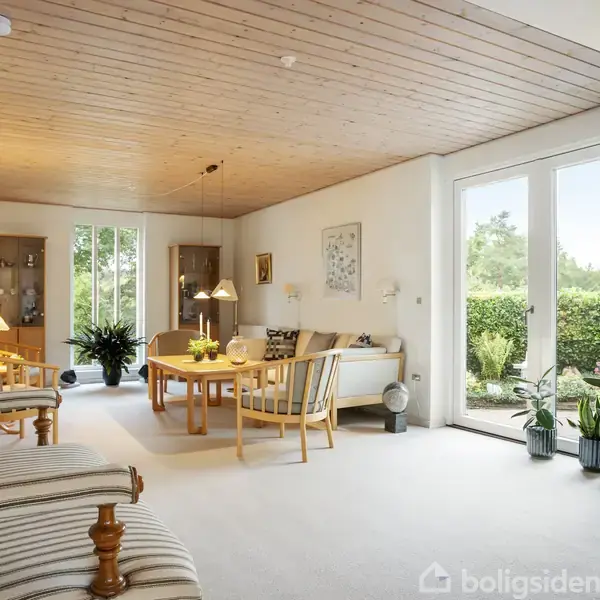 Stue med polstrede møbler står mod træbeklædt loft, og store vinduer åbner op til en frodig have. Planter og lamper dekorerer rummet, skaber en lys og hyggelig atmosfære.