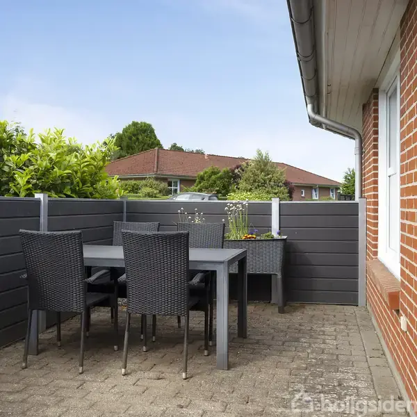 Et moderat stort bord med sorte kurvestole står på en flisebelagt terrasse. Terrassens omgivelser inkluderer en murstensvæg til højre og en høj hæk til venstre.
