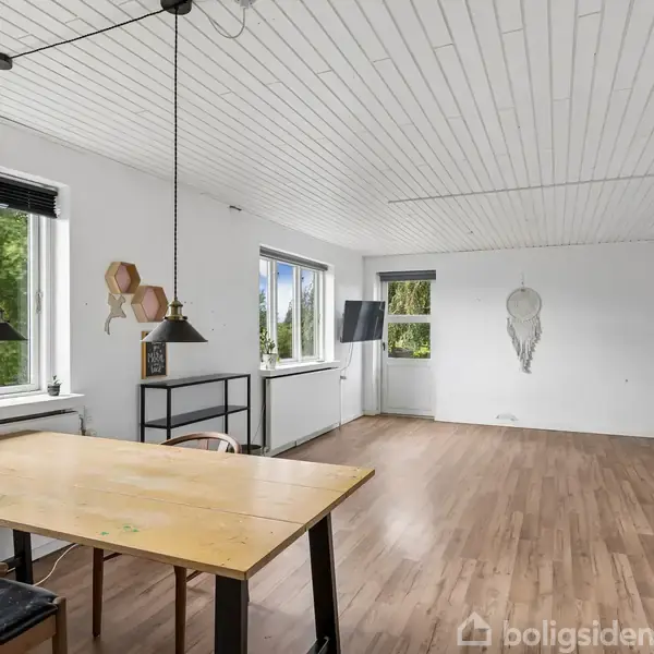 Træbord står i et rummeligt, lyst rum med parketgulv. Ved siden af hænger to sorte lamper fra hvidt loft. Vinduer og glasdør viser frodigt grønt udenfor.