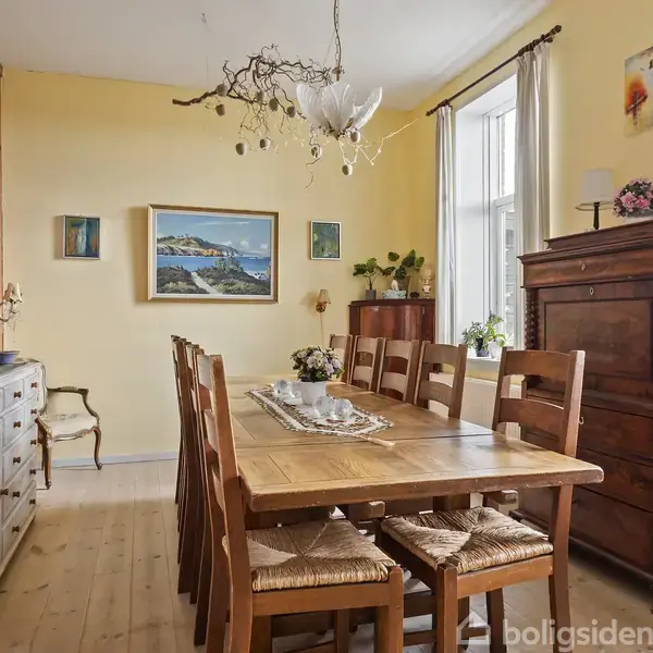 En lang spisebord med træstole står i et gult spisestue. Der er en lysekrone med grene over bordet og malerier på væggen. Gardiner hænger ved to vinduer.