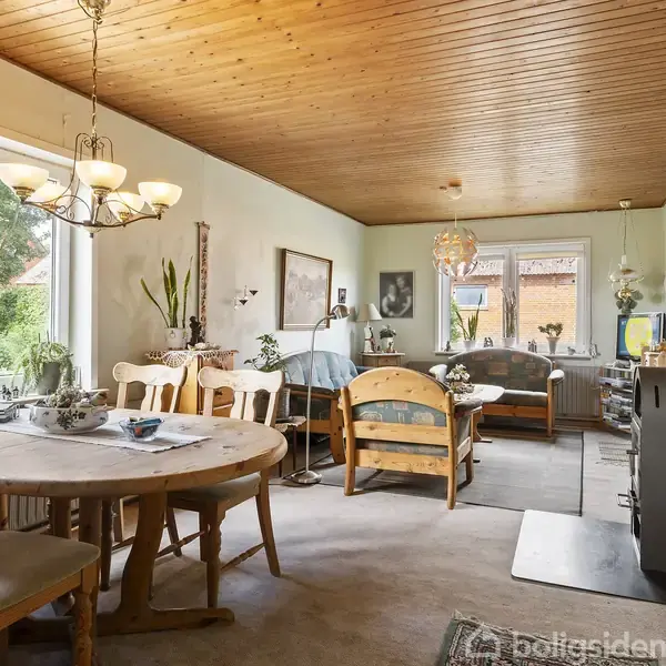 Spisebord står i et lyst rum med træloft, flankeret af stole. I baggrunden ses en sofa og en brændeovn. Store vinduer giver udsigt til en grøn have.