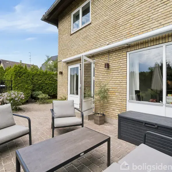 Møbelgruppe med sofa, stole og bord står på en flisebelagt terrasse foran gulstenshus med store vinduer. Omgivelserne inkluderer grønne buske og en blå himmel.