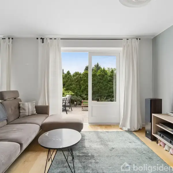 Gråt sofaarrangement står i en lys stue med et stort vinduesparti og glasdør ud til terrasse. En fladskærmstv står på et lavt tv-bord. Lysebeige gardiner hænger ved vinduerne.