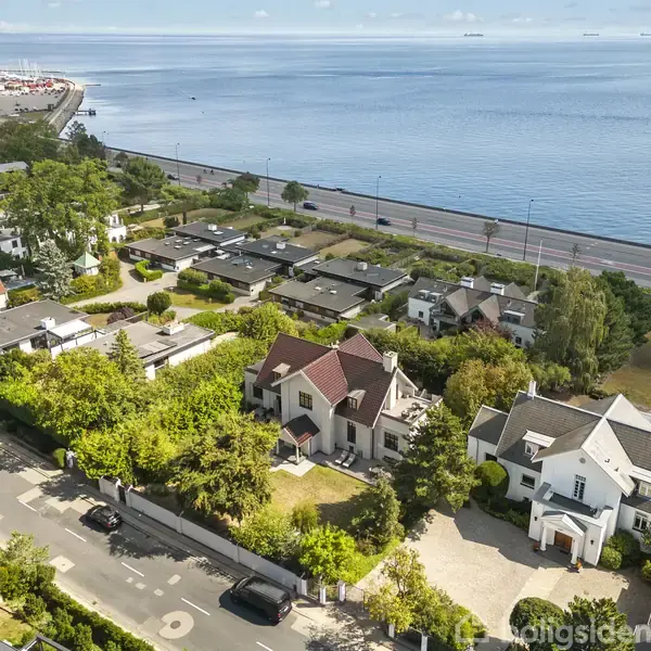 Huse ligger i en frodig grøn kvarter nær en kystlinje. Veje og biler ses i forgrunden, mens havet strækker sig i baggrunden.