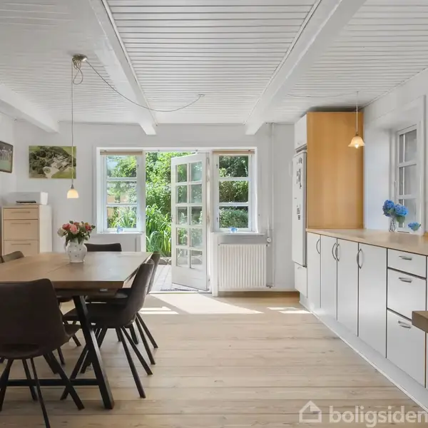 Spisebord med stole står i et lyst køkken. Skydedøre åbner mod en have, og køkkenet er udstyret med hvide skabe og trædetaljer. Blomster og dekorationer pryder rummet.