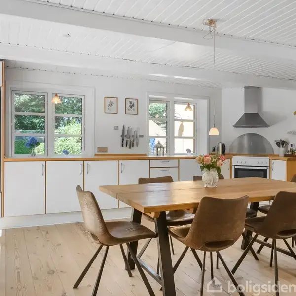 Køkkenbord med seks stole står i et lyst køkken med trægulv og hvide skabe. Et komfur med emhætte i baggrunden. Vinduer giver udsigt til grønne omgivelser. Ingen tekst.